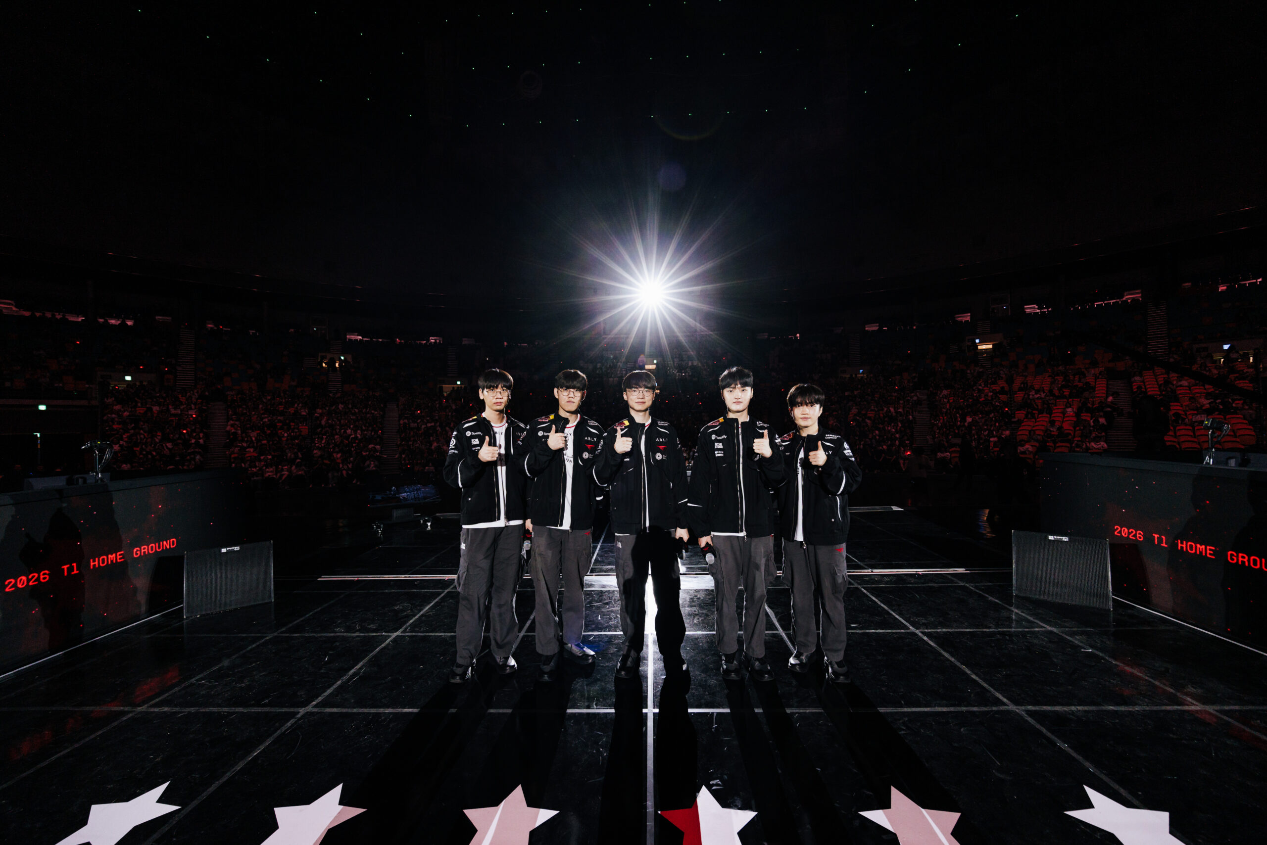 Five T1 esports players in black team jackets stand in a row on a dark arena stage, giving thumbs-up under a bright spotlight. LED boards read "2026 T1 HOME GROUND."